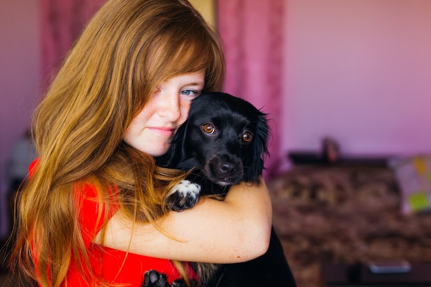 Menina com sardas no rosto, segurando um cachorro preto nos braços e abraços