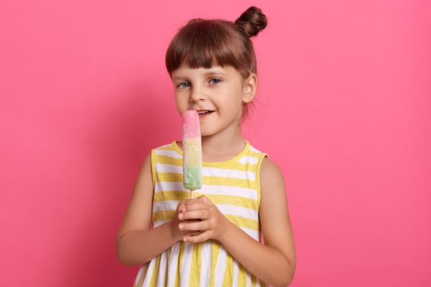 Menina com sorvete, olhando diretamente para a câmera, usando um vestido de verão branco e amarelo, posando contra a parede rosa, lambendo o sorvete, parece satisfeita.