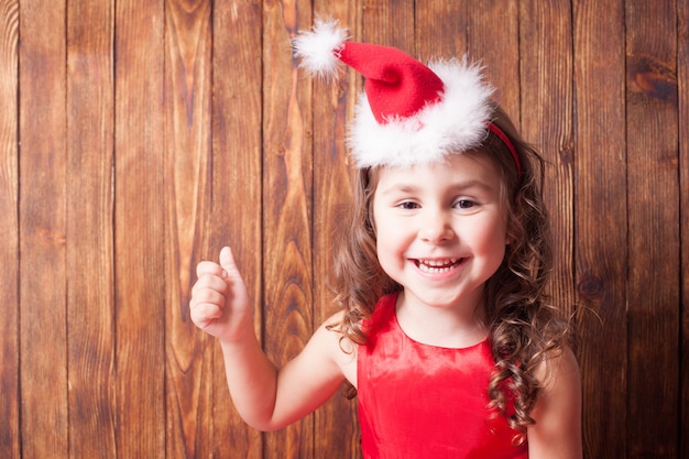 Menina com tiara de chapéu de Papai Noel sentada na parede de madeira e mostrando OK, copie o espaço