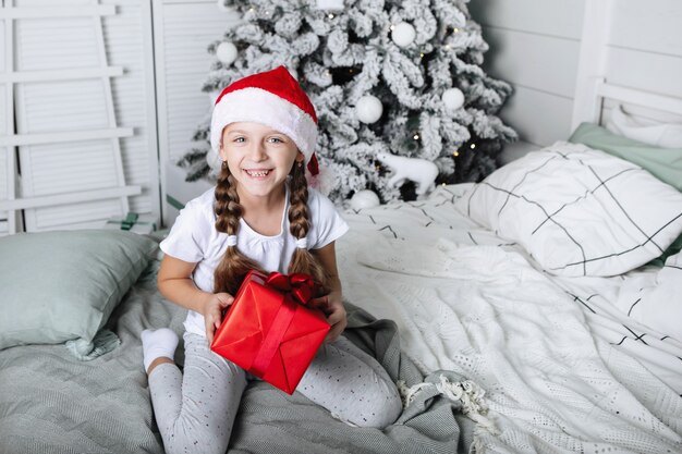 Menina com tranças em um boné vermelho de Natal se senta em uma cama com uma caixa de presente.