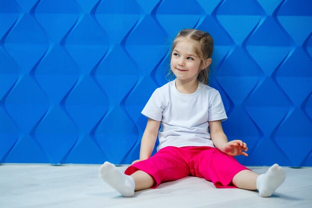 Menina com tranças em uma camiseta branca sobre um fundo azul. Ela se senta em posição de lótus no chão e sorri e acena com as mãos.