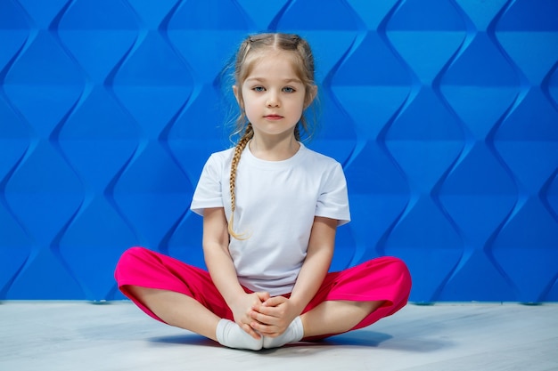 Menina com tranças em uma camiseta branca sobre um fundo azul. Ela se senta em posição de lótus no chão e sorri e acena com as mãos.