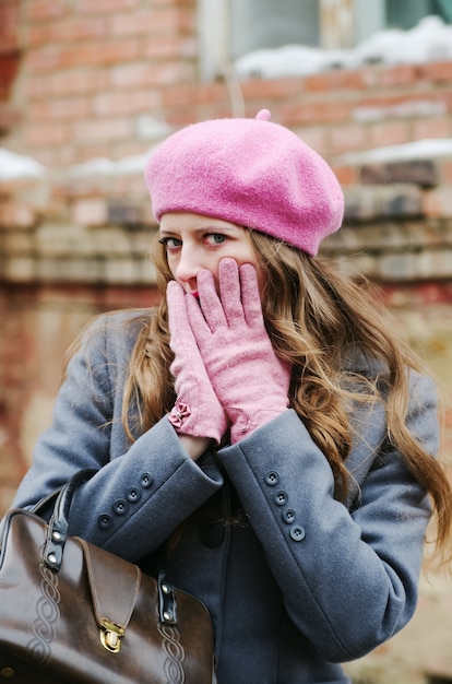 Menina com um casaco cinza e uma boina rosa, cobre o rosto com as mãos