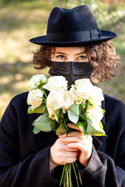 Menina com um casaco preto, chapéu e máscara descartável preta com um buquê de rosas brancas ao ar livre. Dia ensolarado de primavera.