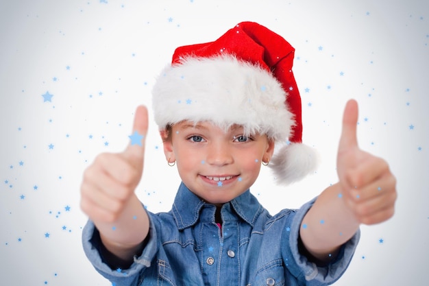 Menina com um chapéu de Natal e os polegares para cima contra a queda de neve