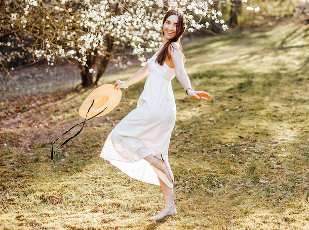 Menina com um chapéu de palha na primavera no parque. Morena com cabelo comprido segura um chapéu em um fundo de natureza de verão. Juventude e beleza.