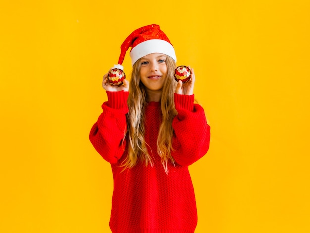 Menina com um chapéu de papai noel e um suéter vermelho salta e mostra cinco em um fundo amarelo.