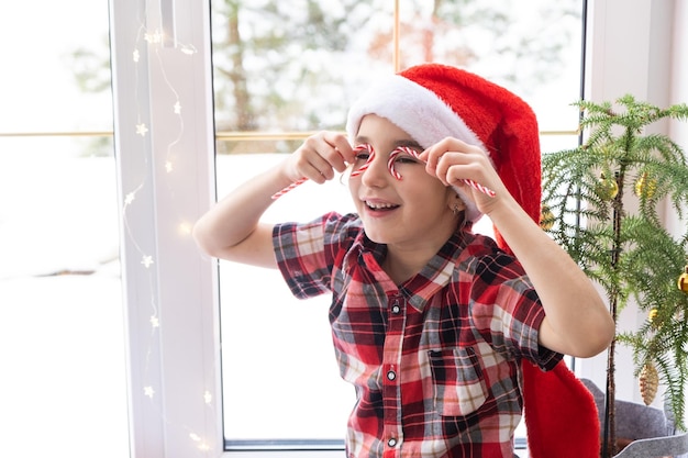 Menina com um chapéu de Papai Noel senta-se no peitoril da janela de uma casa perto da árvore de Natal e coloca bastão de doces nos olhos como óculos A criança está se divertindo e fazendo caretas esperando o Natal e o Ano Novo