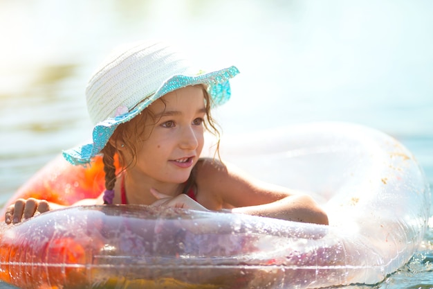 Menina com um chapéu nadando no rio com um círculo inflável transparente em forma de um coração com penas laranja dentro. O mar com fundo de areia. Férias na praia, natação, bronzeamento, protetores solares.