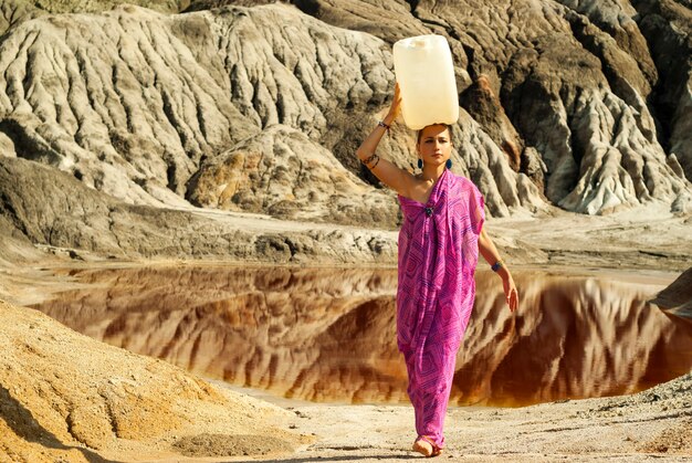 Menina com um sári carrega na cabeça uma grande vasilha de plástico com água sobre uma área deserta