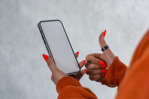 Menina com um suéter laranja com lindas unhas segura uma maquete de smartphone com tela branca nas mãos. tecnologia mock-up.