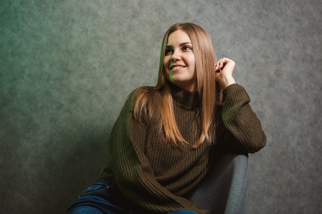 Menina com um suéter verde e calça jeans em uma cadeira cinza e sorrindo