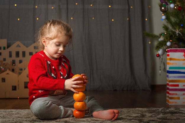 Menina com um suéter vermelho com um veado para o ano novo. Uma menina e tangerinas na árvore de Natal.