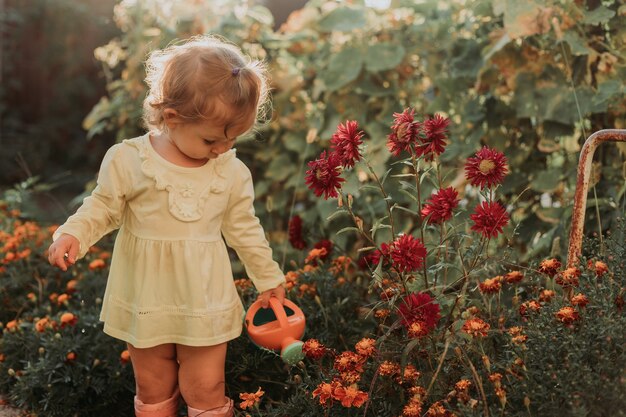 Menina com um vestido amarelo e botas de borracha regando flores no jardim Ajudante