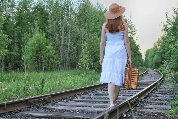 Menina com um vestido de verão branco e uma mala de vime andando sobre os trilhos