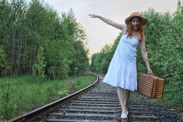 Menina com um vestido de verão branco e uma mala de vime andando sobre os trilhos