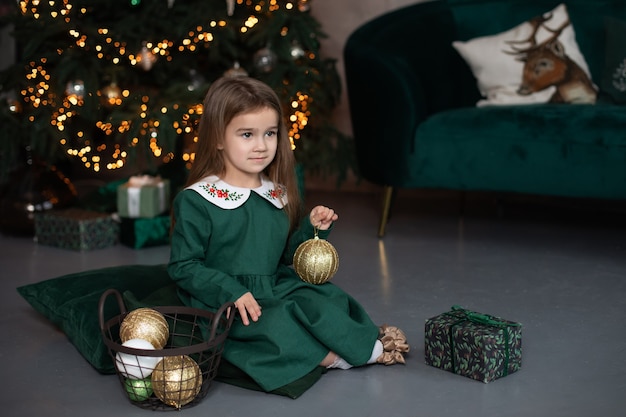 Menina com um vestido verde sentada no chão da sala de estar decorando a árvore de Natal com bolas