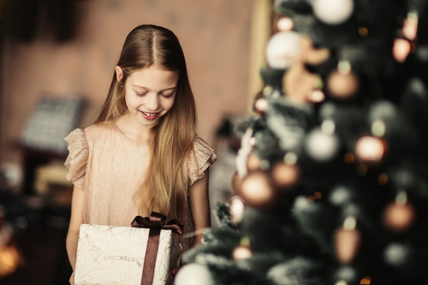 Menina com uma caixa de presente em pé perto da árvore de Natal.