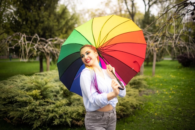 Menina com uma camisa azulada com maquiagem brilhante e longas tranças coloridas. Sorrindo e segurando um guarda-chuva com as cores do arco-íris no fundo de um parque florido, aproveitando a próxima primavera.