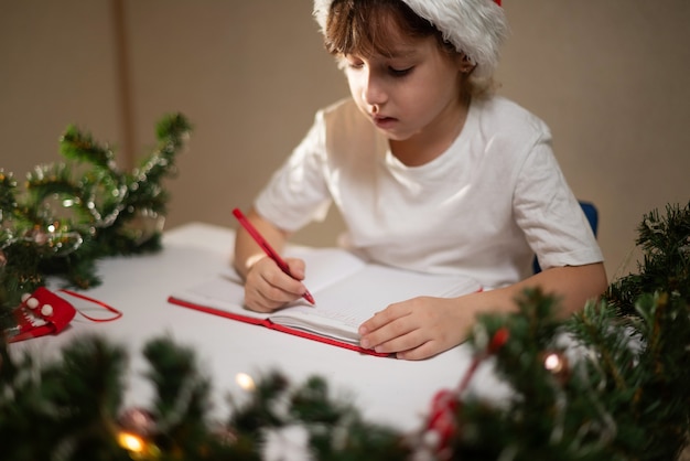 Foto menina com uma camiseta branca com uma caneta vermelha e um boné de ano novo na cabeça escreve uma carta do querido papai noel à mesa em antecipação ao nascimento. lista de desejos de presentes para o ano novo.