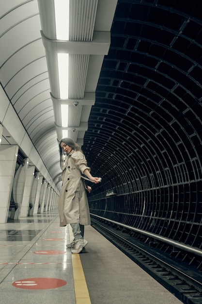 Foto menina com uma capa impermeável bege no metrô esperando o trem na plataforma