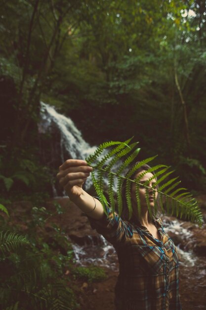 Foto menina com uma folha de samambaia na floresta com uma cachoeira no fundo xa