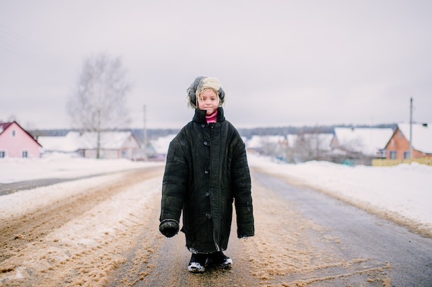 Menina com uma jaqueta grande em uma estrada com neve no inverno