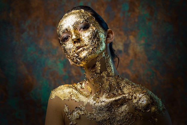 Menina com uma máscara no rosto feita de folha de ouro Retrato de estúdio sombrio de uma morena em um fundo abstrato