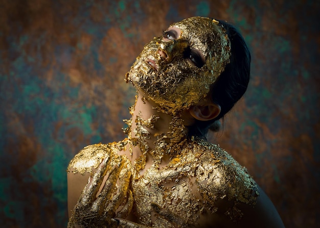 Menina com uma máscara no rosto feita de folha de ouro Retrato de estúdio sombrio de uma morena em um fundo abstrato