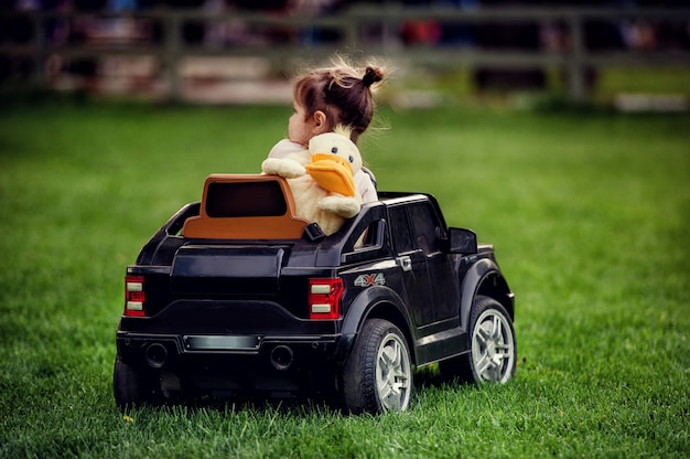 Menina com uma mochila em forma de pato nas costas, dirigindo um carro controlado por rádio grande cabriolet preto na grama verde no parque no verão com fundo desfocado