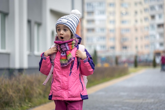 Menina com uma mochila em uma jaqueta e um chapéu perto do espaço da cópia da escola