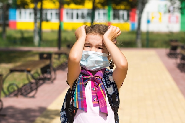 Menina com uma mochila perto da escola depois das aulas tira uma máscara médica, infeliz, cansada e com dor de cabeça. Ele segura a cabeça com as mãos, falta oxigênio. Coronavírus, medidas de segurança