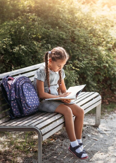 Menina com uma mochila sentada em um banco e lendo um livro perto da escola De volta às aulas de escola agendar um diário com notas Educação aulas do ensino fundamental De volta à escola