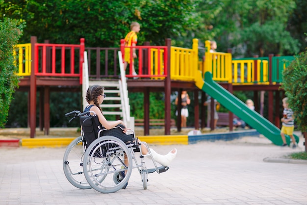 Menina com uma perna quebrada senta-se em uma cadeira de rodas em frente ao parque infantil