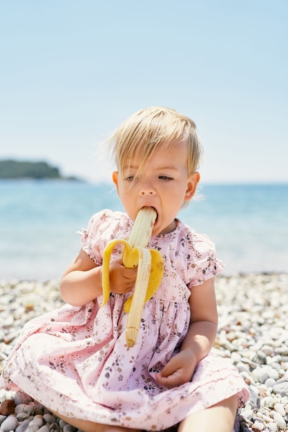 Menina comendo banana sentada em uma praia de seixos