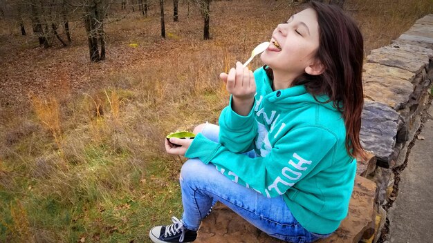 Foto menina comendo comida no campo enquanto está sentada em uma parede de contenção