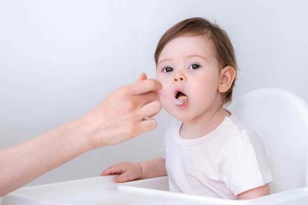 Menina comendo mistura de comida amassada sentada na cadeira alta mãe alimentando a mão da criança com colher para o almoço de vegetais bebê desmame primeiro alimento sólido para criança