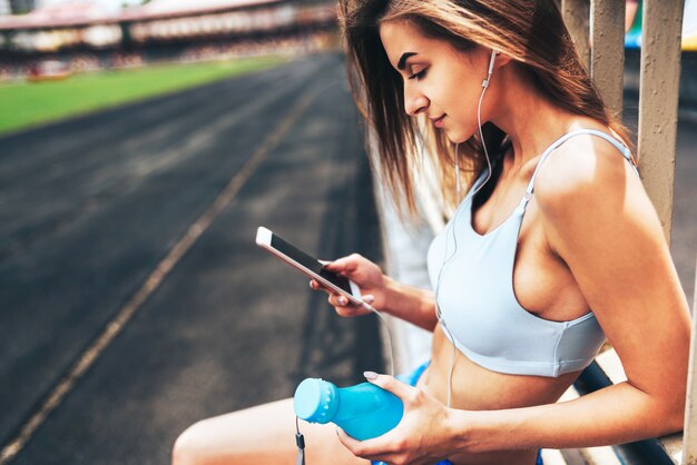 Menina consideravelmente desportiva que relaxa e que escuta a música no telefone após o treinamento