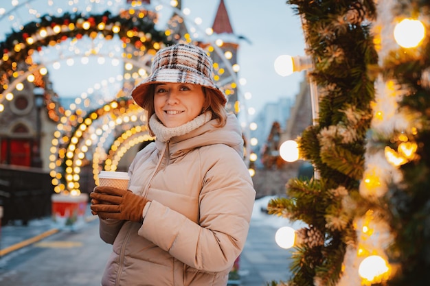 Menina copo bebida quente enquanto caminhava no mercado de Natal decorado com luzes do feriado à noite. feliz na cidade grande.