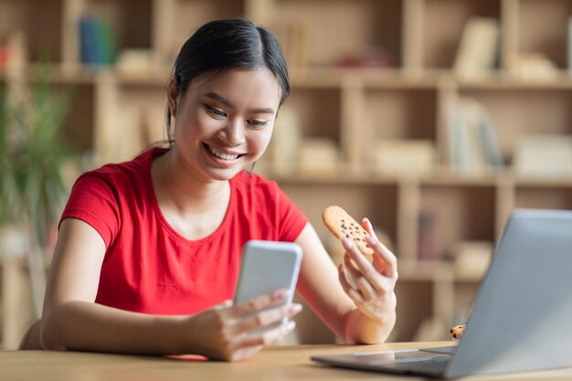 Menina coreana adolescente satisfeita olha para o telefone segurar o cookie na mesa com o pc no interior da sala