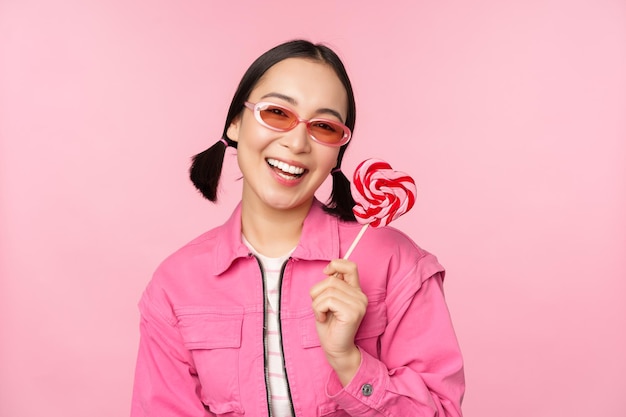 Menina coreana estilosa lambendo pirulito comendo doces e sorrindo de pé em óculos de sol contra fundo rosa