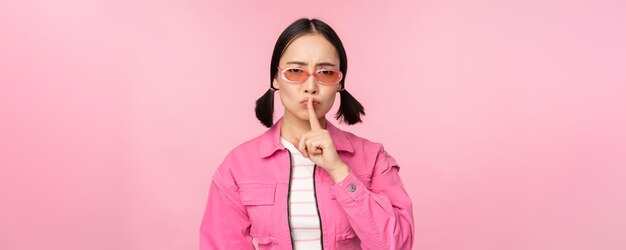 Menina coreana moderna em óculos de sol elegantes de primavera mostrando sinal de silêncio pressione o dedo nos lábios gesto tabu em pé sobre fundo rosa