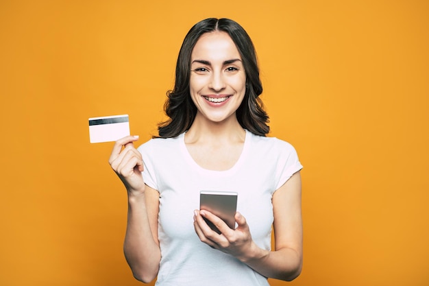 Menina corpulenta com olhos castanhos, cabelo levemente cacheado e um sorriso tão brilhante na frente de um fundo laranja extravagante segurando seu telefone e um cartão de banco