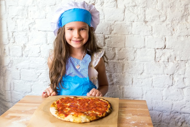 Menina cozinheira sorrindo perto de pizza de pepperoni em forma de coração após a preparação.