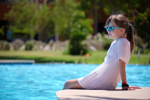 Menina criança alegre no vestido branco descansando no lado da piscina com água azul clara no dia ensolarado de verão Conceito de férias tropicais