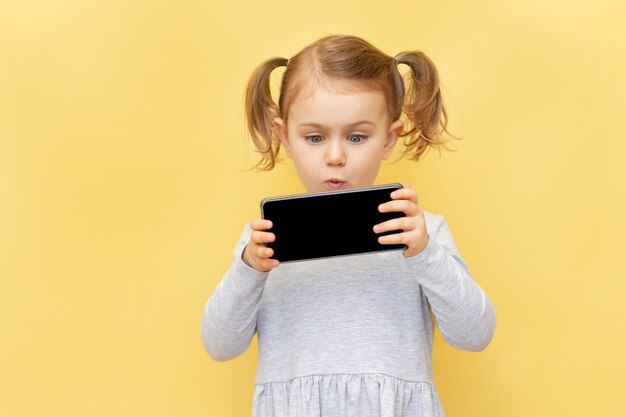 Menina criança chocada segurando um telefone e com expressão de surpresa olha para o telefone na parede amarela