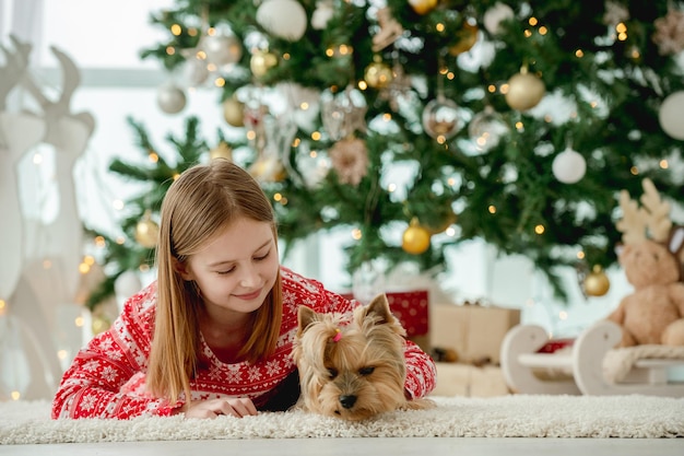 Menina criança com cachorro deitado no chão com árvore de Natal no fundo. Cachorrinho de estimação aproveitando o ano novo em casa