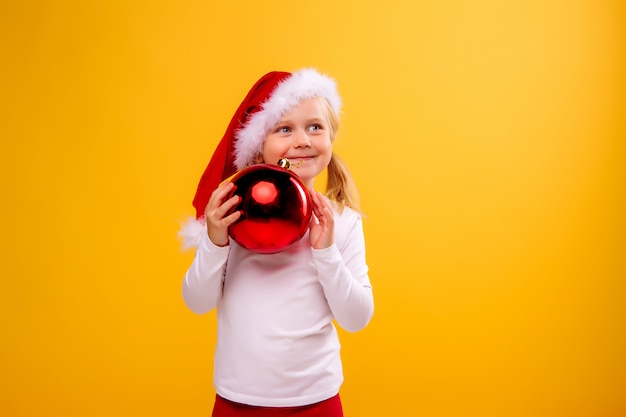 menina criança com chapéu de Papai Noel segurando uma bola de Natal em uma parede amarela
