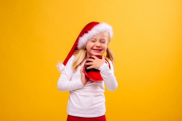 menina criança com chapéu de Papai Noel segurando uma bola de Natal em uma parede amarela