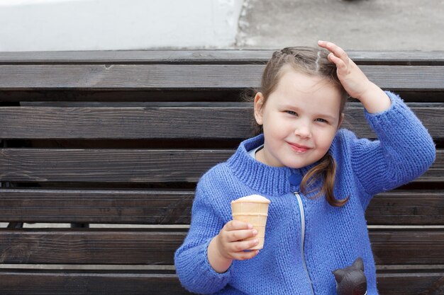 Menina criança com um suéter de malha azul come sorvete sentado em um banco.